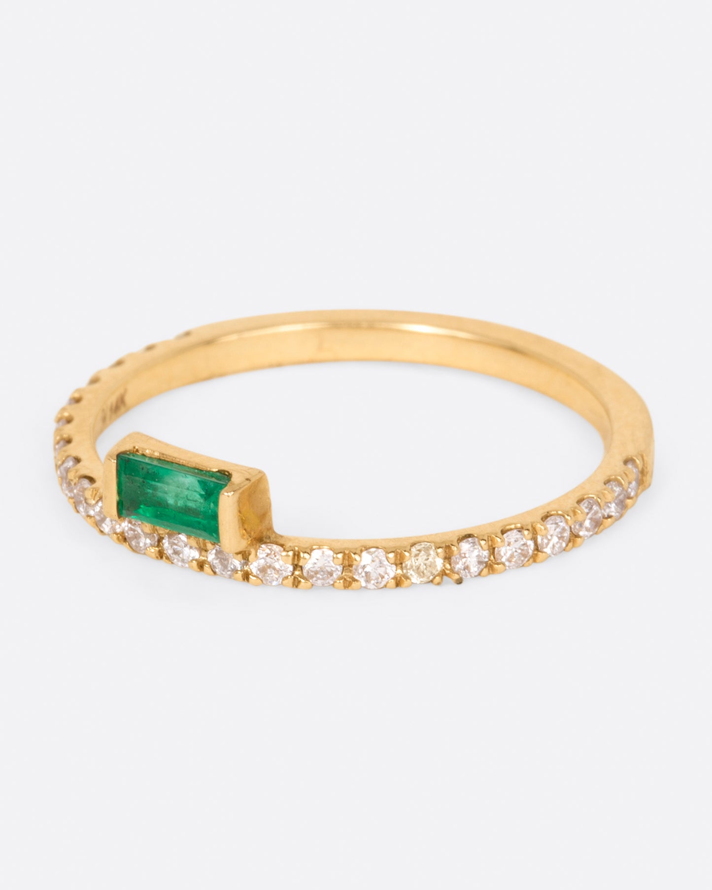 A yellow gold ring with white diamonds going 2/3 of the way around and an emerald baguette sitting atop it, shown from the side.