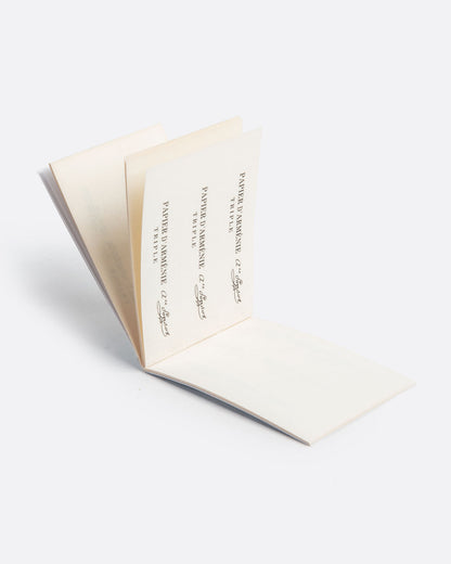 An open booklet of paper incense. Each page has strips of white paper with writing on top. Shown lying flat from the side at an angle.