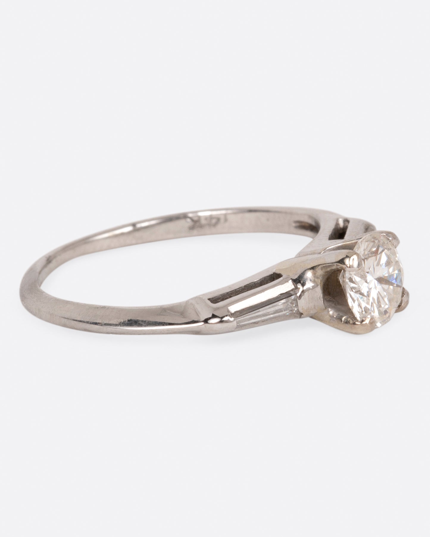 A white gold ring with a round prong-set diamond at the center and tapered baguette diamonds on either side, shown from the side.