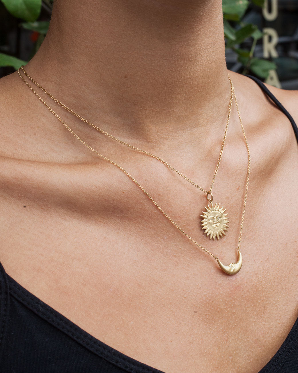 Womans chest wearing two necklaces. The bottom necklace is a tiny yellow gold crescent moon, with a face carved into it and little diamond eyes, is a unique pendant. Double sided and hanging from a chain. The chain is attached to the ends of the crescent, so it falls on a sideways/horizontal angle across the chest.