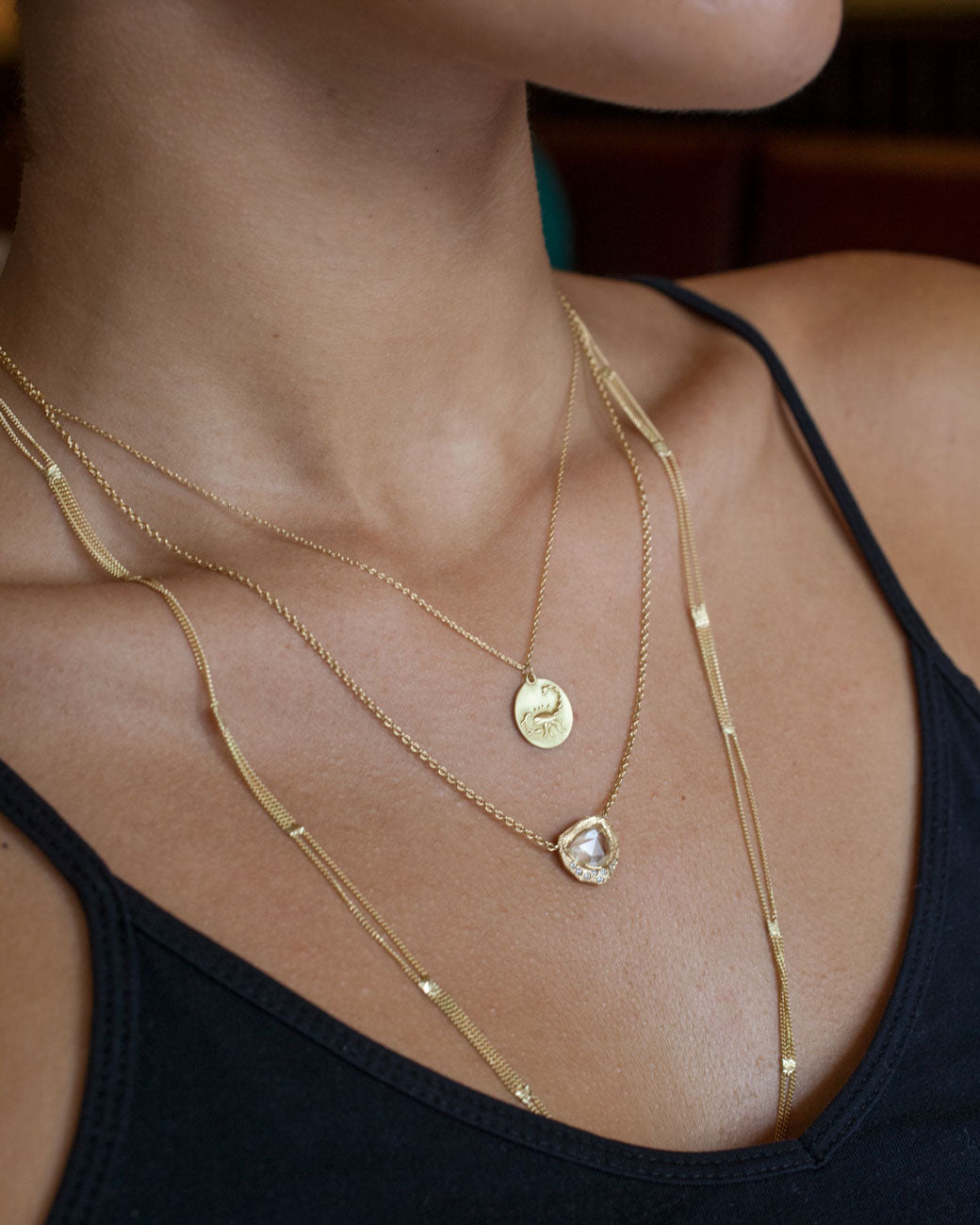 A woman wears three necklaces; one is a gold disc pendant with a Scorpio zodiac design, one is a large white sapphire, and the other is a long multi-chain gold piece. She wears a black dress.